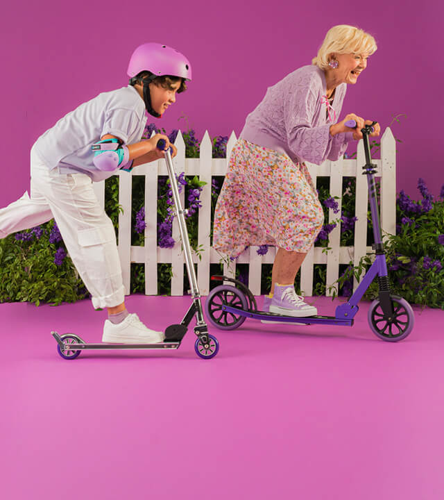 Two people wearing helmets ride kick scooters past a white fence with flowers, against a pink background.