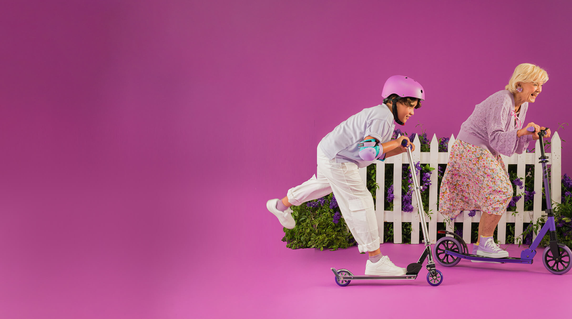 Two people wearing helmets ride kick scooters past a white fence with flowers, against a pink background.