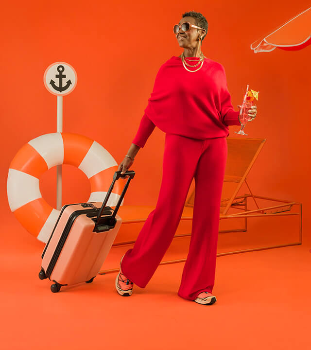 Person in red outfit with sunglasses holds a suitcase and a drink, standing before an orange background with a lifesaver, anchor sign, and lounger.