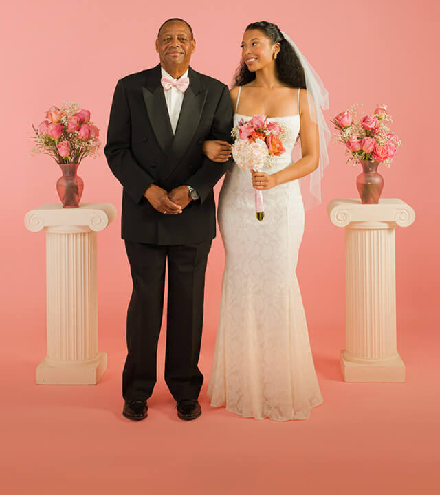 Older man and bride stand joyfully against a pink background. The woman wears a white lace gown and veil, holding a bouquet; the man wears a formal black suit.