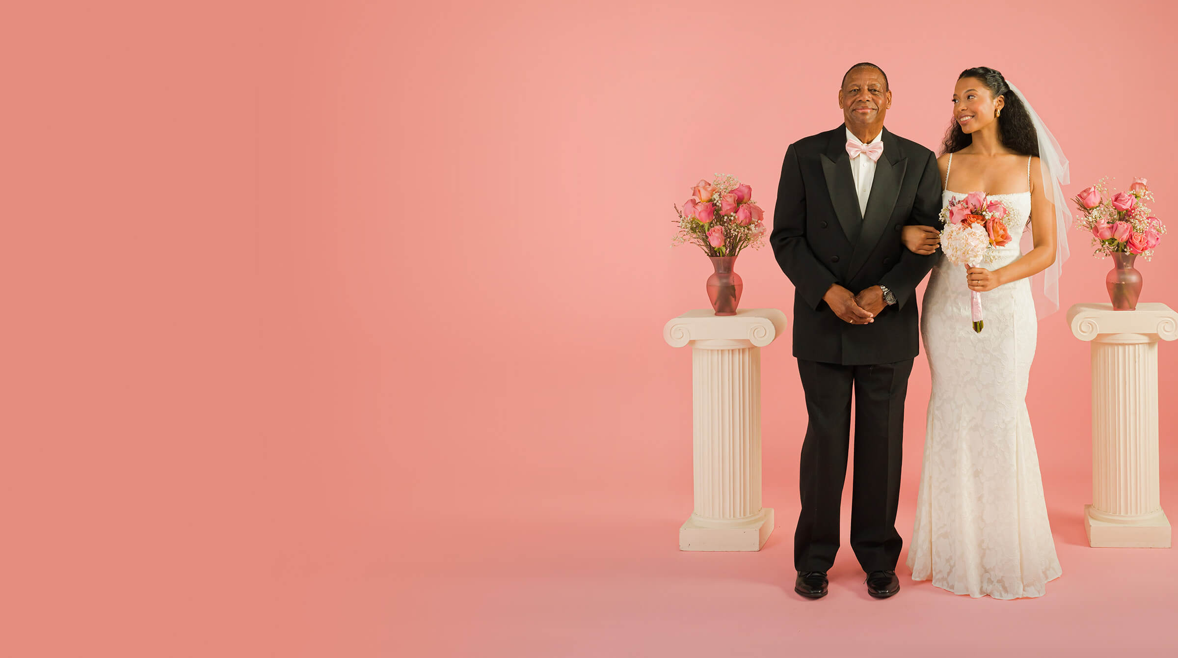 Older man and bride stand joyfully against a pink background. The woman wears a white lace gown and veil, holding a bouquet; the man wears a formal black suit.